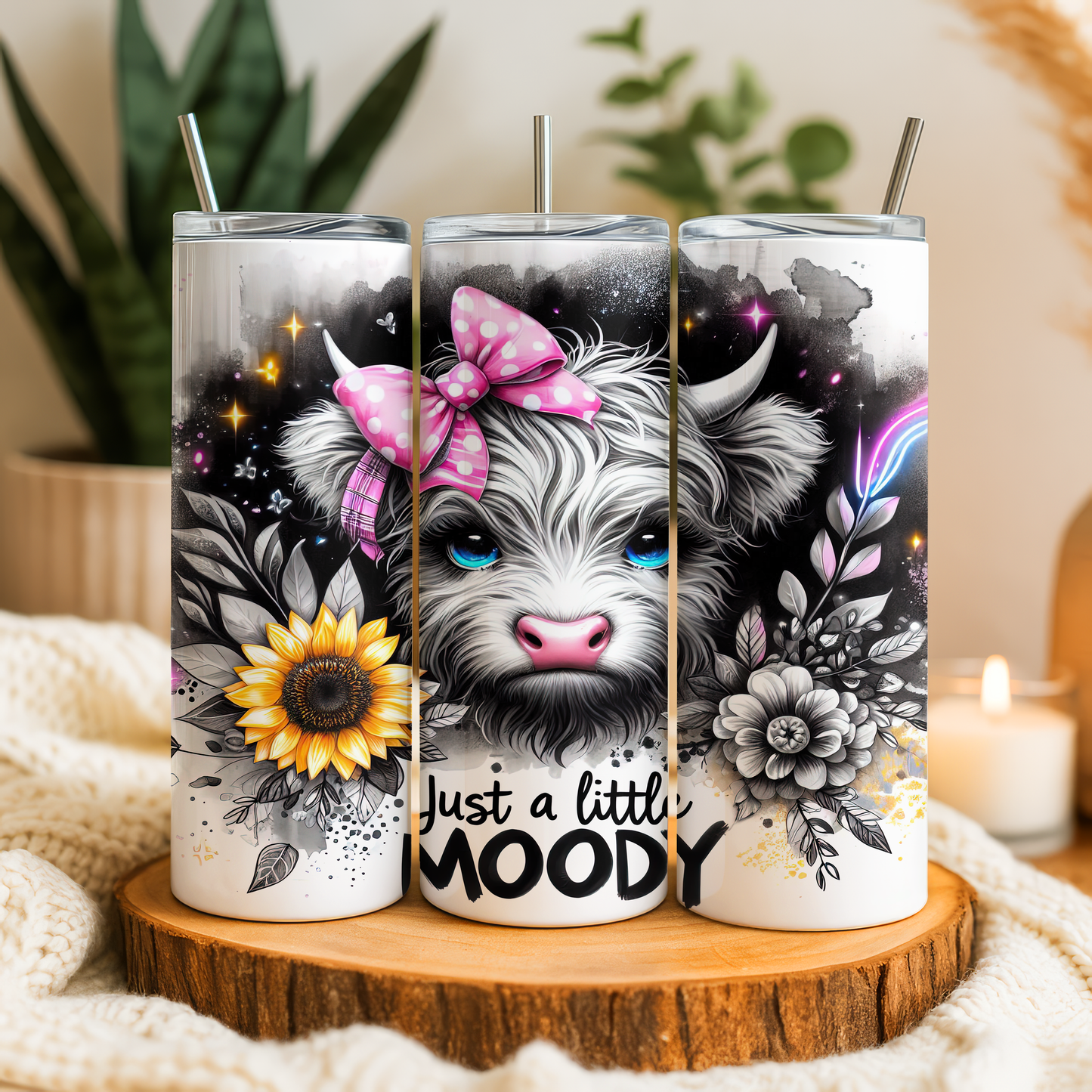 A trio of decorative tumblers with a whimsical design featuring a cow's face with a pink bow, flowers, and the text "Just a little moody".