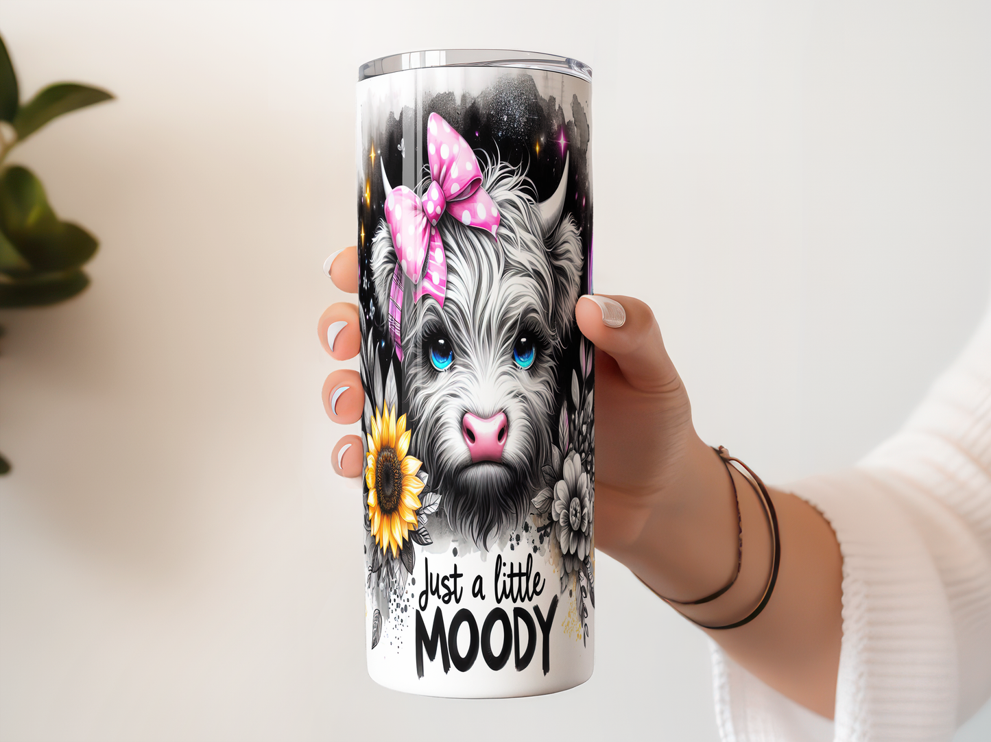 A person's hand holding a white tumbler with a colorful design featuring a cow's face, flowers, and a pink bow.